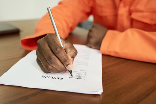 Black man in orange uniform sitting at desk writing on resume document with pen, showing hand and arm, participating in educational or vocational training program in prison setting