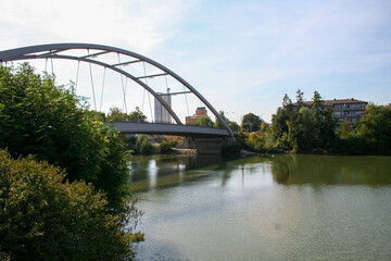 A contemporary arch bridge spans a calm river, connecting parts of a city with buildings and lush...