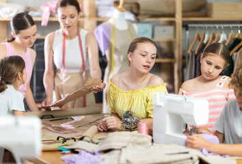 Curious teenage girl attentively watching peer stitching at machine during practical clothing construction course for school students led by experienced sewing instructor in training workshop..