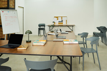 Empty classroom with several laptops, notebooks, and pens arranged on long tables, chairs...