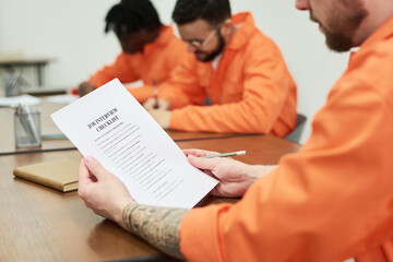 Three young adult men wearing orange uniforms sitting at table studying documents in classroom...