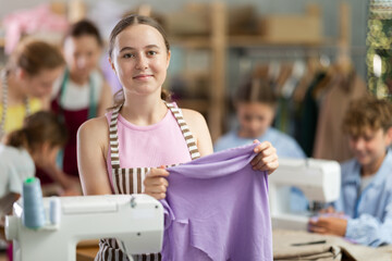 Enthusiastic teenage girl holding sewing work with smile during practical clothing construction...