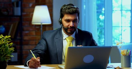 Taking notes in video call on laptop at office desk, Indian Asian businessman with headphones focused on virtual meeting, corporate executive engaged in remote work, digital communication - Powered by Adobe