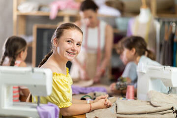 Smiling teenage girl sitting at table during school sewing workshop, while friendly, experienced...