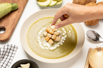 Hand adding croutons to Zucchini, onion and potato soup served on beige dish
