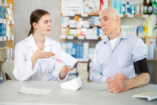 Young female pharmacist offering pills to mature man having blood pressure measured
