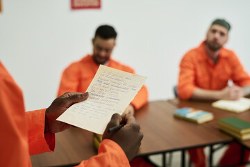 Black man holding handwritten letter standing in foreground while two men in orange uniforms...