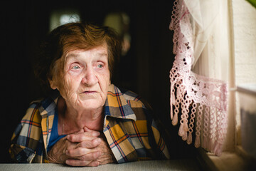 An elderly woman, wrapped in a man's shirt draped over her shoulders, gazes thoughtfully out the...