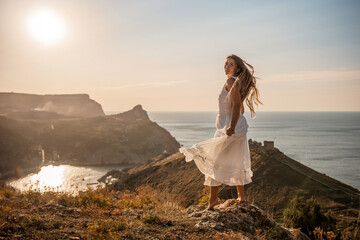 A woman stands on a hill overlooking the ocean. She is wearing a white dress and has long hair. The...
