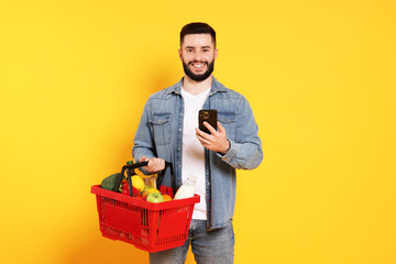 Man with shopping basket full of products and smartphone on orange background
