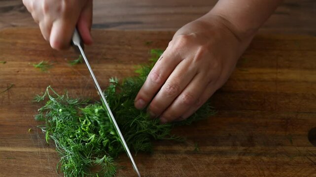 Female hands cutting fresh dill on wooden cutting board in kitchen, close-up. Concept of organic food, healthy eating, homemade cooking, food preparation for winter.