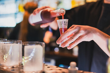 Bartender pouring red liquor into jigger at bar counter, with ice-filled glasses in foreground. Close-up of cocktail preparation in nightlife setting