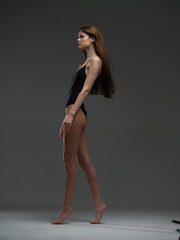 Fototapeta premium slender woman posing in black bodysuit with long brown hair emotionally expressing elegance on gray studio background, full body side view in soft lighting