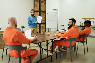 Middle aged Caucasian man instructing three multiethnic male prisoners wearing orange jumpsuits...