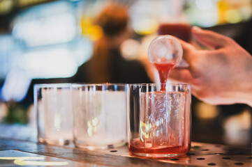 Bartender pouring a vibrant red cocktail into a glass with ice cubes at a bar counter, close-up...