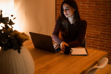 Young woman working late at home with warm lamp light – student or freelancer using laptop for...
