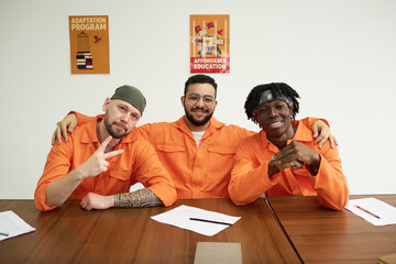 Three young adult inmates sitting at table wearing orange uniforms, smiling and posing together,...