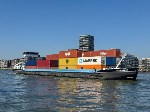 Dordrecht, Netherlands - 1 May 2025: Colourful inland container barge sails by Dordrecht on the calm Maas River in the Netherlands. Stacked metal containers reflect the efficient transport of freight