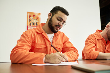 Young adult man wearing orange uniform sitting at table writing on paper with pen, focused...