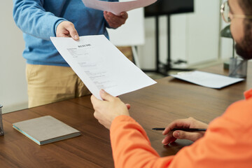 Young man sitting at desk receiving printed resume from standing middle aged man during educational...