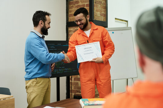 Middle aged man congratulating young adult man in orange prison uniform receiving certificate during educational program in classroom setting with another inmate observing