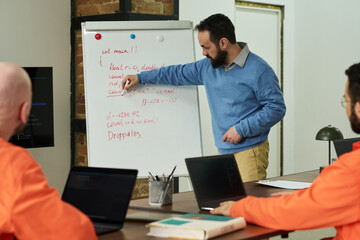 Middle aged Caucasian man explaining programming concepts on whiteboard to two Black men in orange uniforms sitting at table with laptops, during prison education session