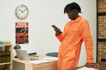 Black teenage boy wearing orange jumpsuit standing in office setting, leaning on desk and using...