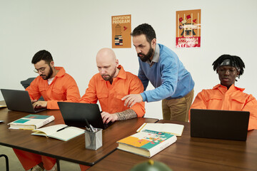 Middle aged Caucasian man instructing group of adult men working on laptops in classroom setting,...