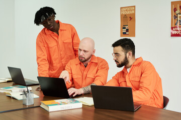 Three young adult men collaborating at table using laptops and books, discussing project in...