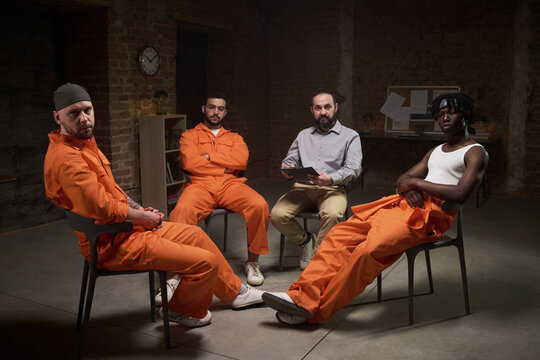 Group of young male convicts sitting in circle participating in group therapy session in prison setting with therapist holding clipboard leading discussion