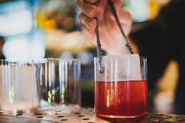 Bartender placing a large ice cube into a glass with red cocktail using tongs, close-up on hand and drink, blurred background, bar counter, modern nightlife concept.
