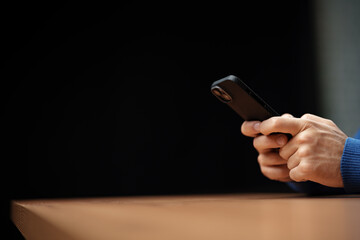 Person using a smartphone while sitting at a wooden table in a dimly lit indoor space during...