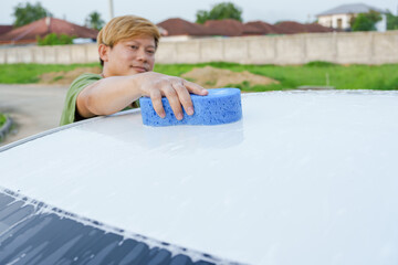 A smiling man is washing his car with soapy water under bright sunlight, showing his enthusiasm and joy in his daily work.