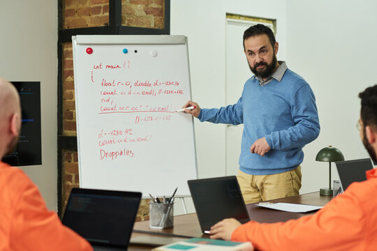 Middle aged Caucasian man teaching programming to group of adult men in orange uniforms in classroom setting, standing by whiteboard with handwritten code, students listening attentively