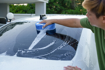 A smiling man is washing his car with soapy water under bright sunlight, showing his enthusiasm and joy in his daily work.
