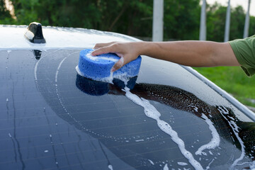 A smiling man is washing his car with soapy water under bright sunlight, showing his enthusiasm and joy in his daily work.