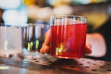 Close-up of a hand holding a glass with red cocktail and ice cubes, with blurred bar background....
