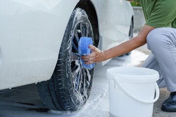 A smiling man is washing his car with soapy water under bright sunlight, showing his enthusiasm and joy in his daily work.