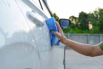 A smiling man is washing his car with soapy water under bright sunlight, showing his enthusiasm and joy in his daily work.