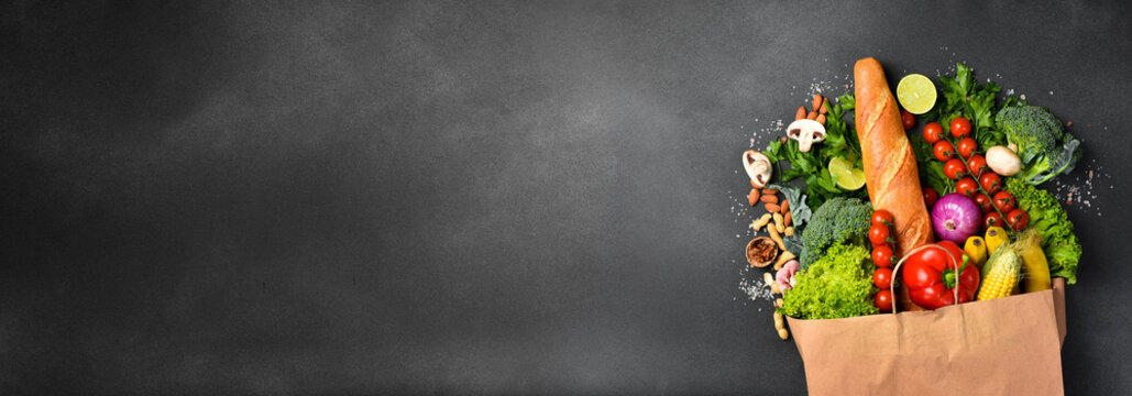 Food background. Wide banner with paper bag filled with fresh vegetables, fruits and various purchases from the market. Free space for text or design. On a black background.