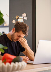 Tired man sitting at desk with laptop in kitchen. Fatigued person rubbing his eyes while working at...