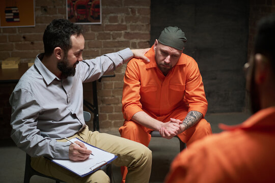 Caucasian man in orange prison uniform sitting with head down being comforted by middle aged Caucasian man holding clipboard during counseling session in prison