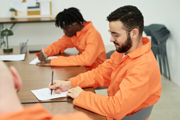 Two diverse male convicts sitting at table writing on paper in classroom setting, participating in...
