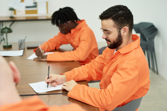 Two diverse male convicts sitting at table writing on paper in classroom setting, participating in educational program inside prison environment