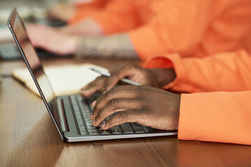 Black man wearing orange uniform typing on laptop during educational program in prison classroom, hands visible on keyboard, other inmates studying in background, focus on learning process