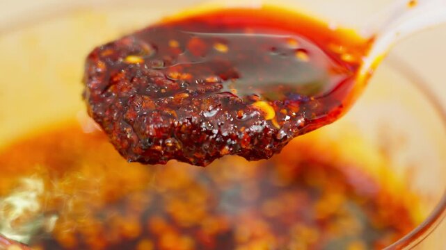 A spoon lifts clumped chili oil packed with chili flakes and seeds&mdash;capturing sticky texture, bold red color, and the rustic intensity of artisanal Chinese spicy condiment.