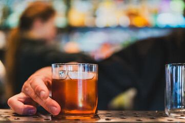 Hand holding a glass of iced whiskey on a bar counter, with a warm, blurred background of colorful...