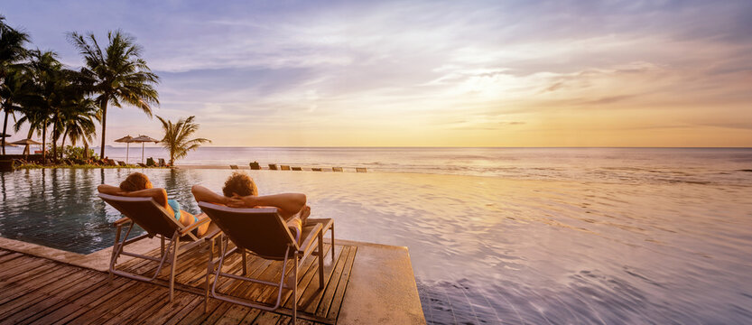 beach vacations, young couple in luxury hotel resort, man and woman relaxing near swimming pool at sunset