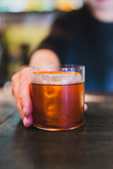 Close-up of a hand holding a glass of amber cocktail with ice and orange peel garnish, on a dark wooden table with a blurred background.