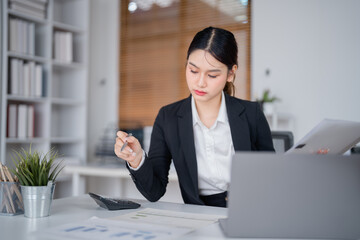 Young businesswoman sitting at her desk and analyzing financial data using laptop and documents, holding a pen and thinking about strategy, working with concentration in modern office
